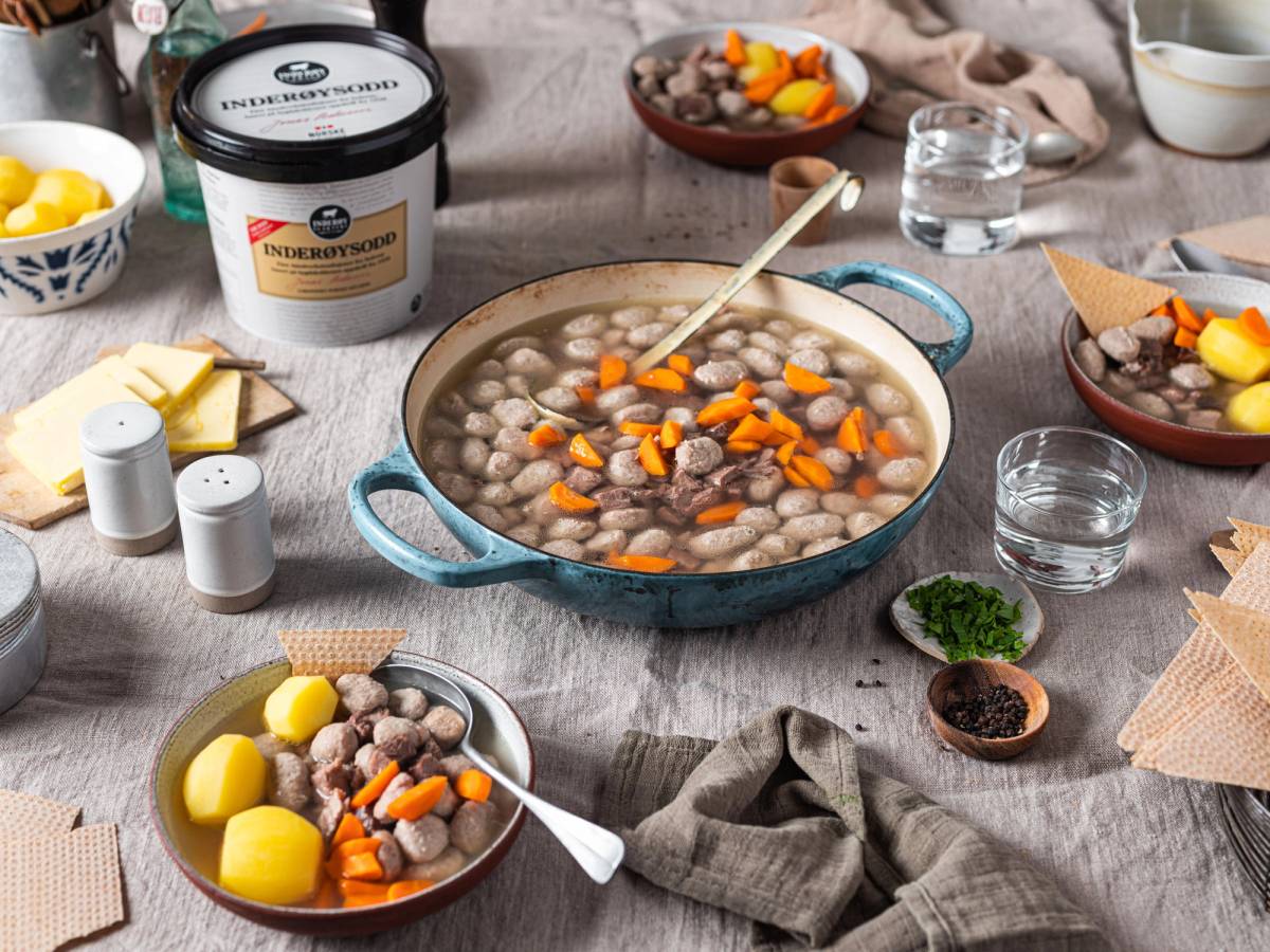 En stor gryte med indisk sodd med kikerter og gulrøtter, omgitt av ulike små boller med tilbehør og krydder på et kjøkkenbord.