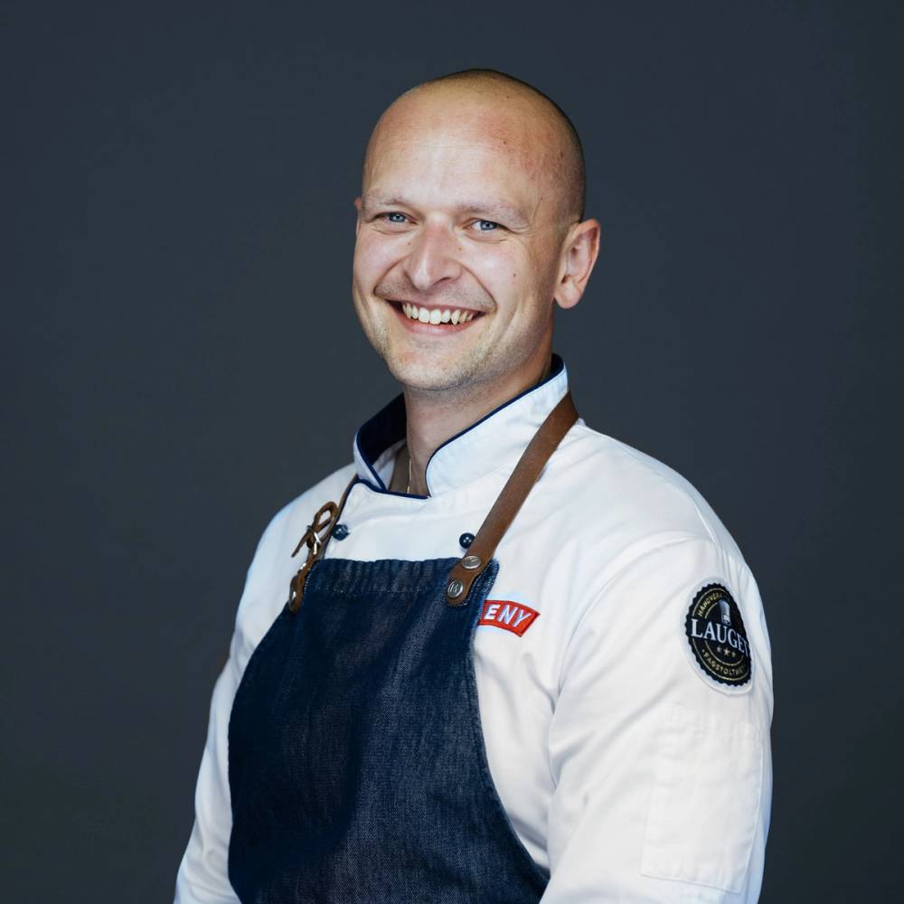 En person iført kokkeuniform og forkle, med en mørk bakgrunn, uten å vise ansiktet.