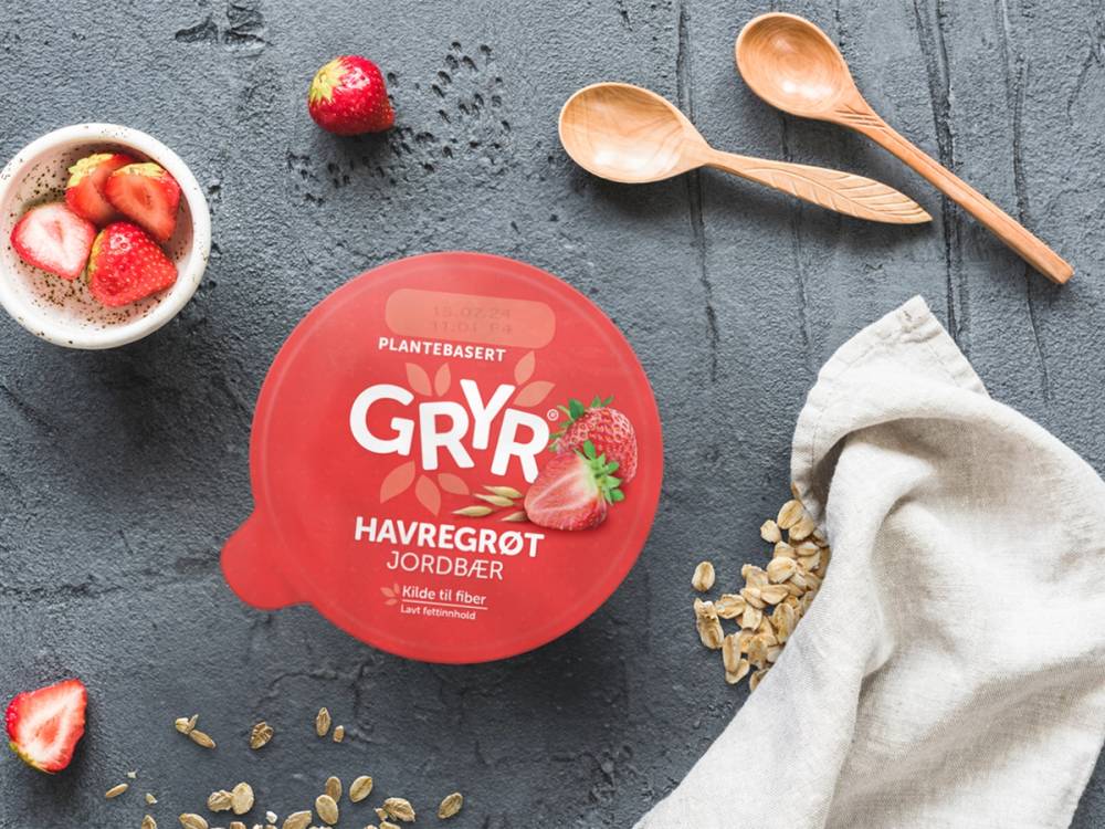 En kopp med jordbær og en pakke med havregrøt fra Gryr på en mørk overflate, med to treskjeer og noen jordbær og havregryn rundt.