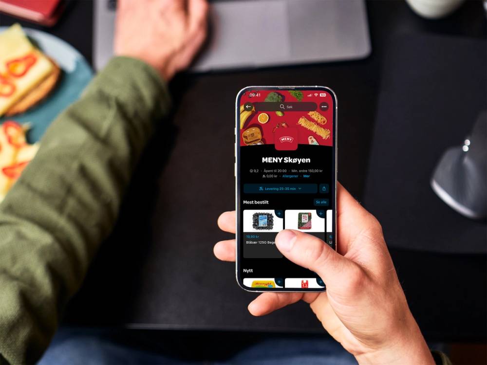 Person som bruker Wolt-appen på mobilen med menyvalg på skjermen, sett fra hånden og overflaten foran.