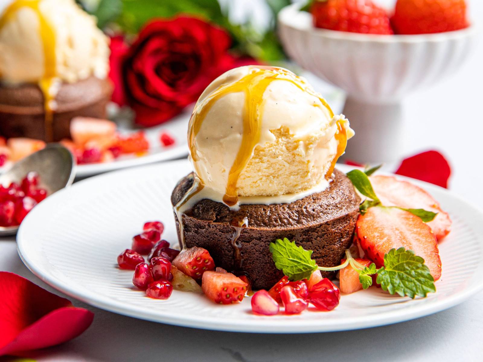 Iskrem med vanilje og karamellsaus servert på en tallerken med friske bær og mynteblader, omgitt av røde roser og andre desserter i bakgrunnen.