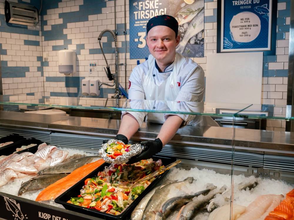 En person i hvit kokkeforkle serverer sushi på en sushi-restaurant, med fisk og sjømat i bakgrunnen.