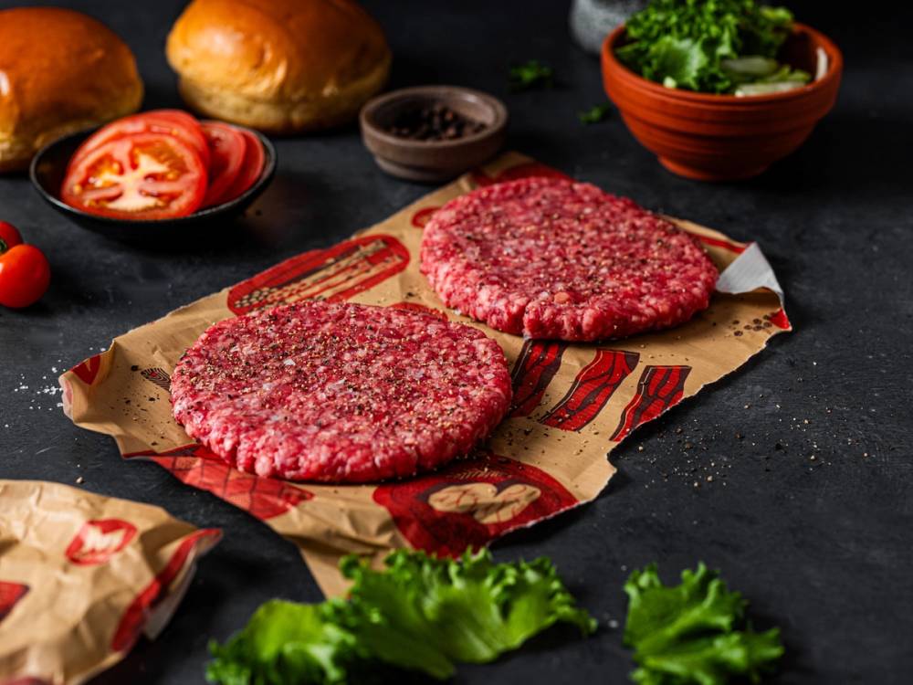 To rå hamburgerbiffer på et stykke brunt papir med grønnsaker og brød i bakgrunnen.