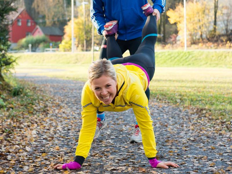 En person i en gul jakke trener yoga utendørs på en grusvei med høstfarger i bakgrunnen.