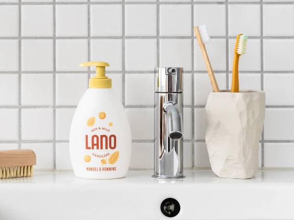 To tannbørster i en hvit keramikkholder og en flaske med LANO håndsåpe på et bademøbel mot en hvit flisvegg