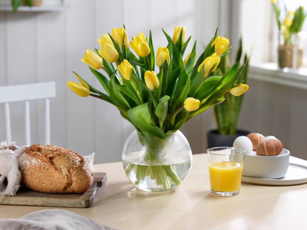 En vase med gule tulipaner på et bord, med frokostmat som brød, egg og juice i bakgrunnen.