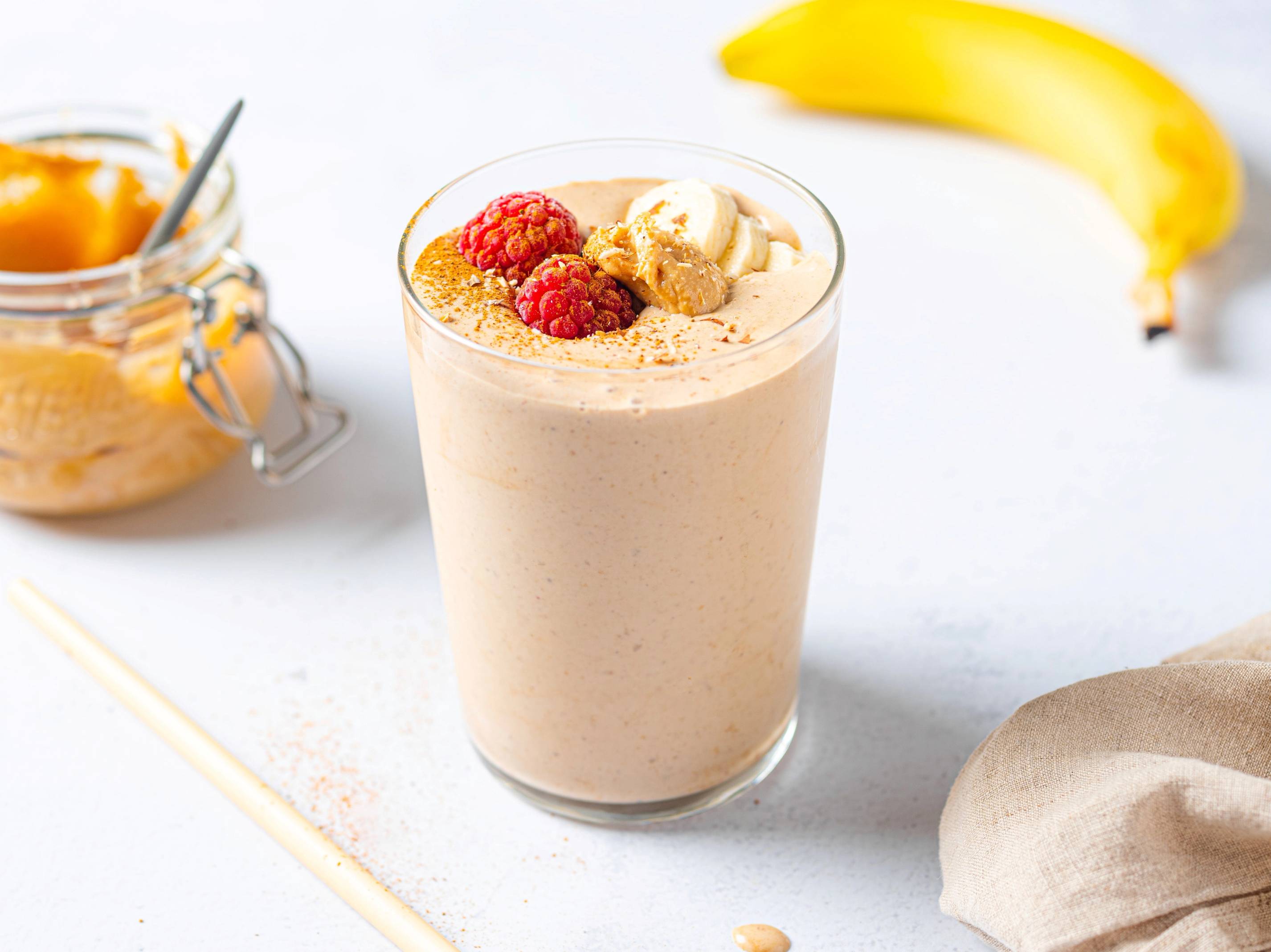 En smoothie med nøttesmør i et glass, pyntet med bær og en skje, med en banan i bakgrunnen.