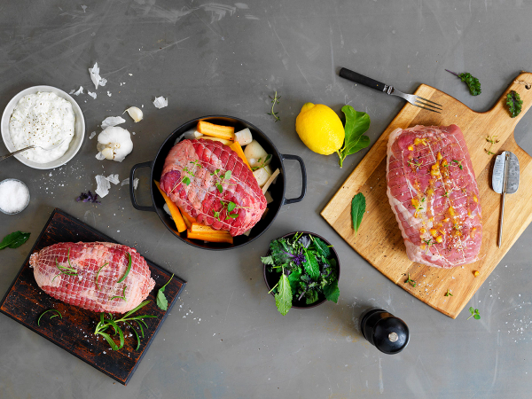Ulike kjøttstykker og urter for å lage en perfekt stek, med tilbehør og kjøkkenutstyr rundt på en mørk arbeidsflate.