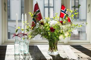 En blomsteroppsats med norske flagg og røde roser på et bord foran et vindu, sannsynligvis til 17. mai-feiring.
