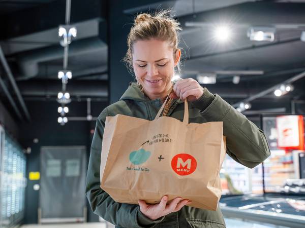Person som holder en papirpose med Too-good-to-go-logo i en butikk eller matbutikk, uten å vise ansiktet.