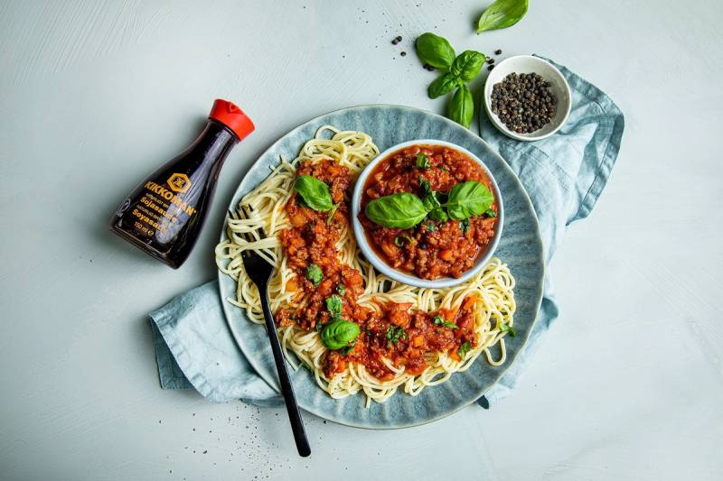 Pasta med kjøttsaus servert på en tallerken med basilikumblader og en flaske Kikkoman soyasaus ved siden av.