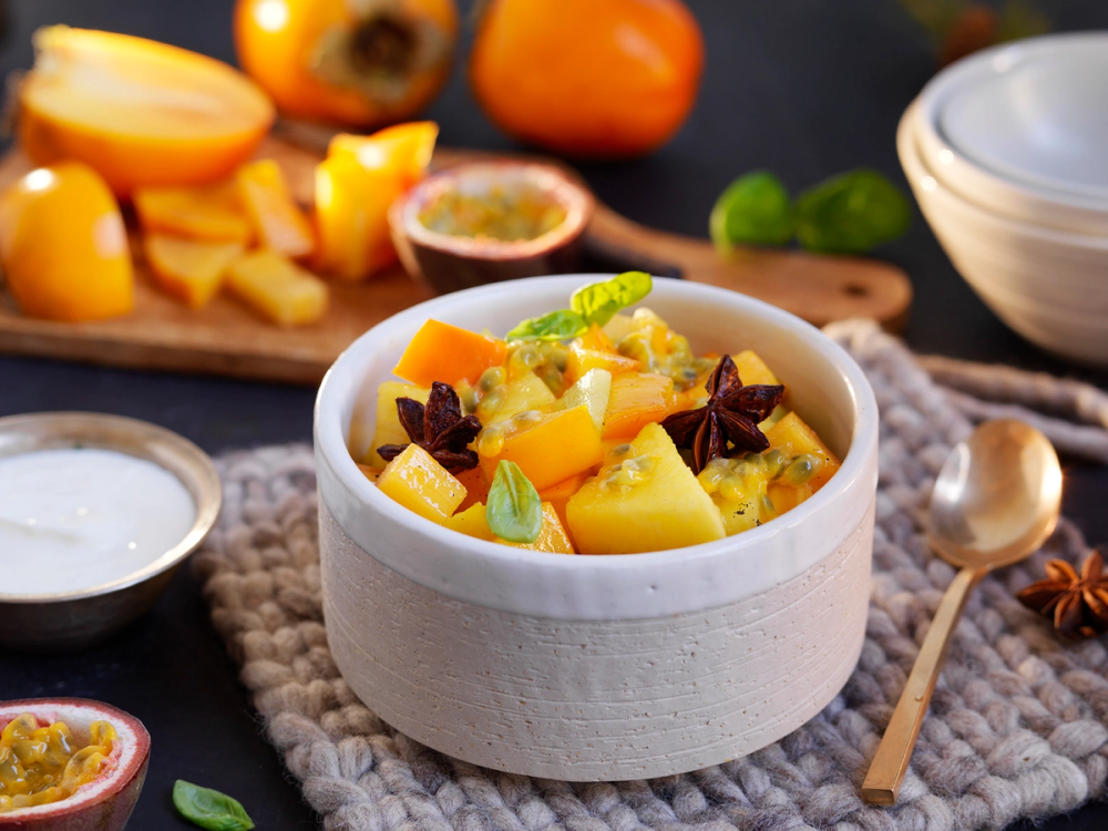Fruktsalat med persimon og vaniljekesam i en hvit bolle på et bord med ulike frukter og en skje ved siden av.