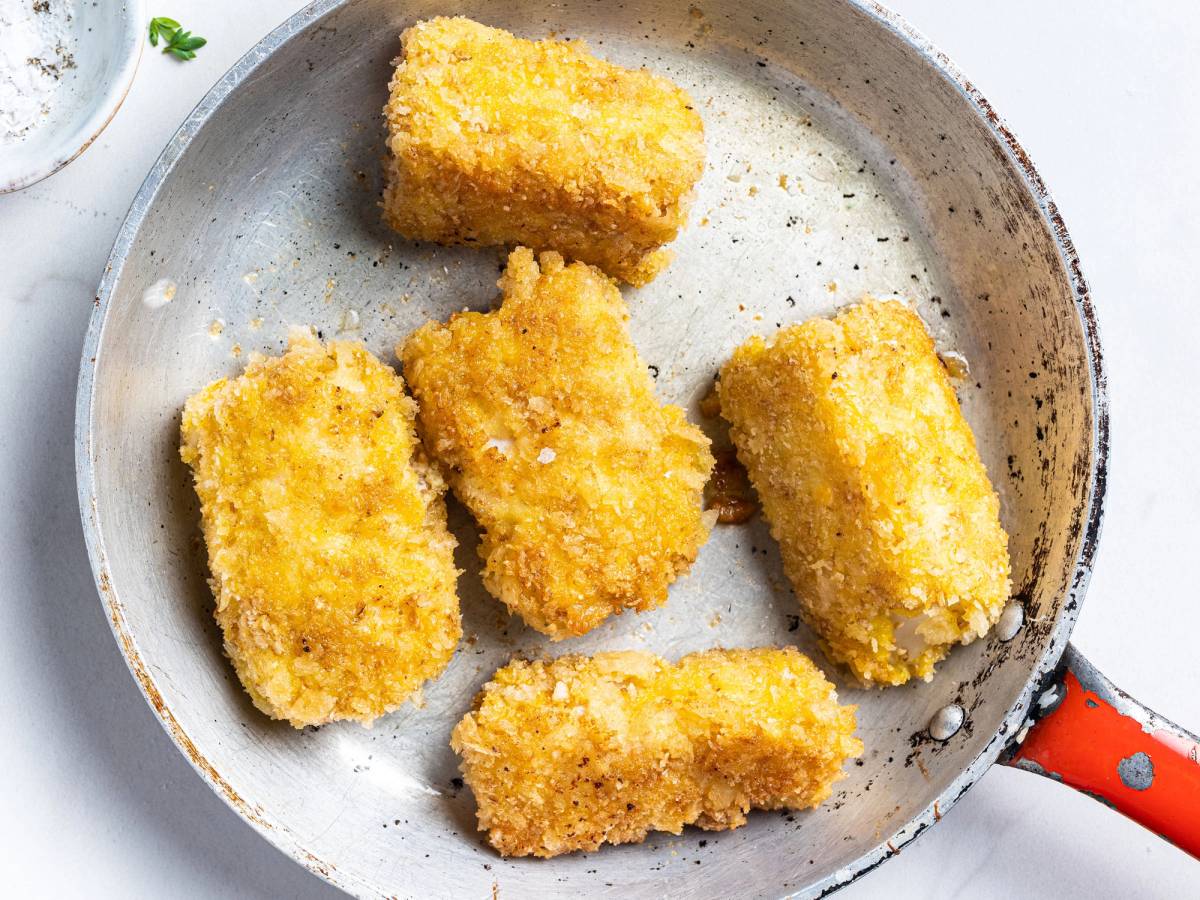 Fem stykker panerte stekebiter i en stekepanne med en rød håndtak, klar til å stekes videre eller serveres.