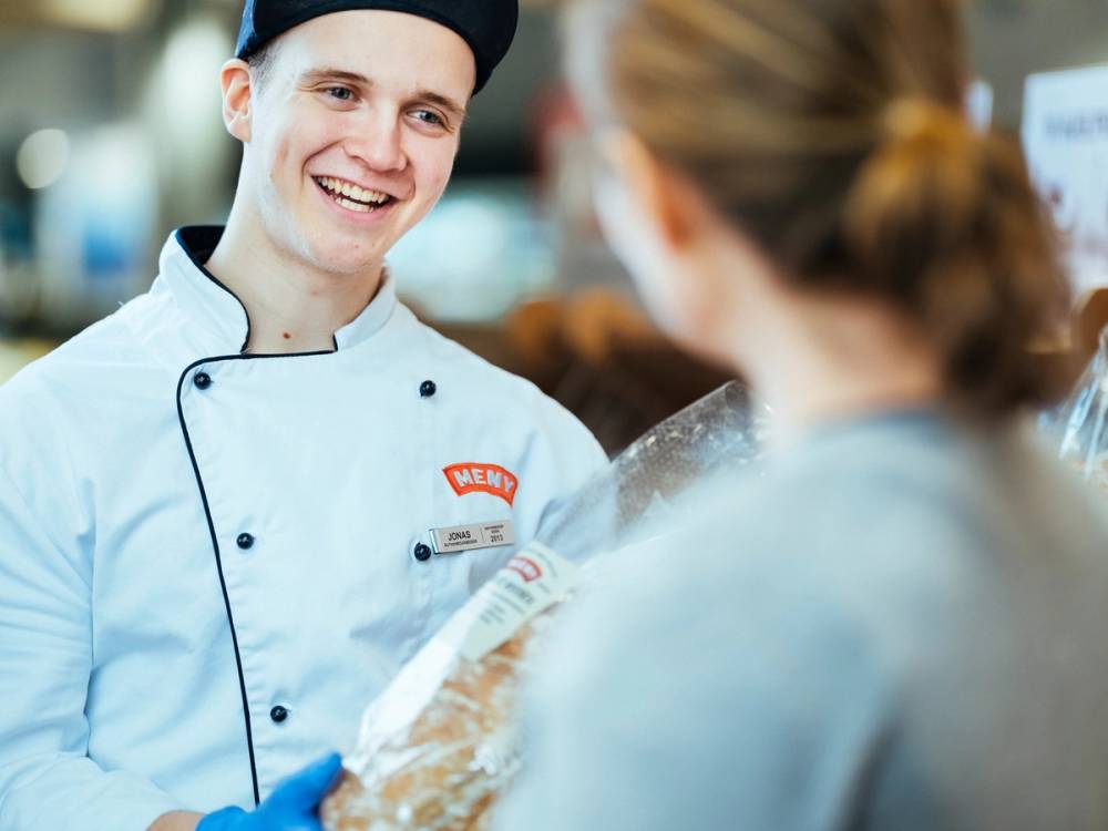 En person i kokkeflate snakker med en kunde i en butikk eller kafé, med en annen kunde i bakgrunnen.