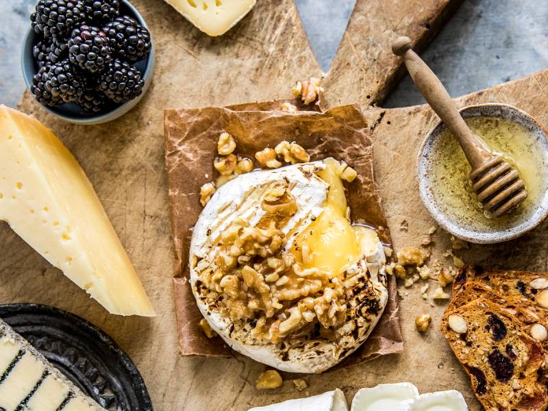 Camembertost med valnøtter og honning på et trebrett, omgitt av ost, bær og kjeks.