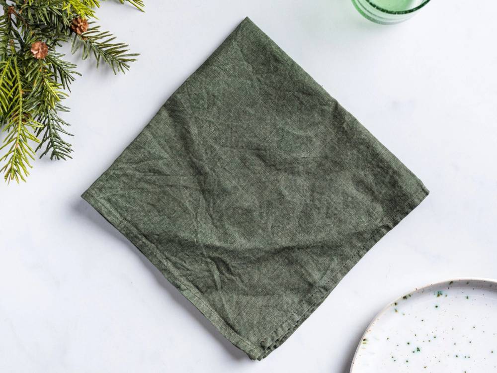 En grønn serviett brettet i form av et juletre på et hvitt bord med noen grønne grener i hjørnet.