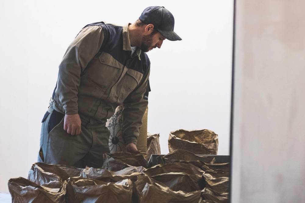 Møllaren Thomas står og ser på flere poser med mat eller korn på et bord, inne i et rom med hvite vegger.