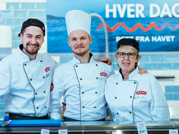 Tre ansatte i fiskedisken iført hvite kokkejakker og chefhatter står bak disken med sjømat, med en blå bakgrunn og en plakat som sier 'Fersk fisk hver dag'.