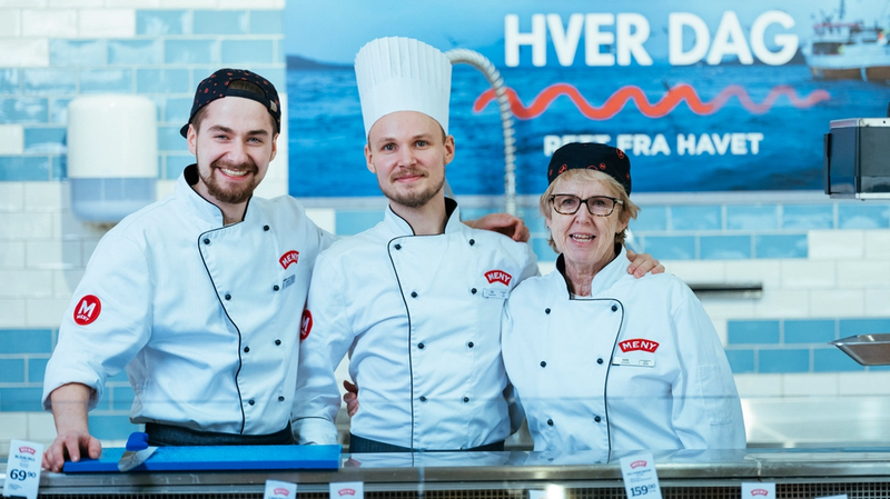Tre ansatte i fiskedisken iført hvite kokkejakker og chefhatter står bak disken med sjømat, med en blå bakgrunn og en plakat som sier 'Fersk fisk hver dag'.