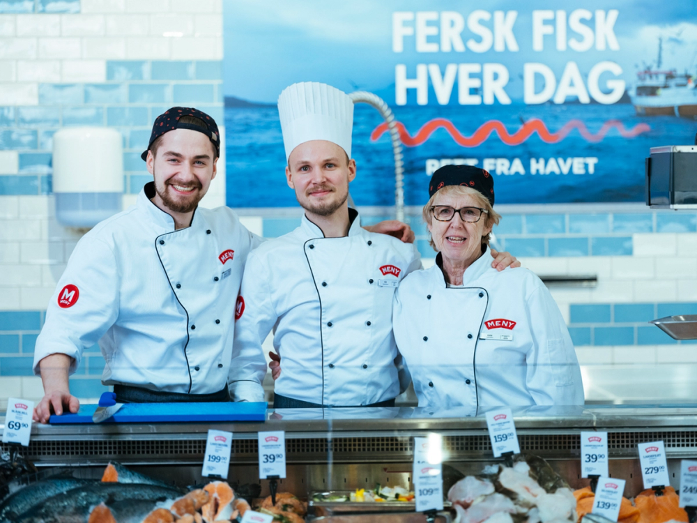 Tre ansatte i fiskedisken iført hvite kokkejakker og chefhatter står bak disken med sjømat, med en blå bakgrunn og en plakat som sier 'Fersk fisk hver dag'.
