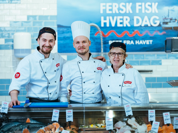 Tre ansatte i fiskedisken iført hvite kokkejakker og chefhatter står bak disken med sjømat, med en blå bakgrunn og en plakat som sier 'Fersk fisk hver dag'.