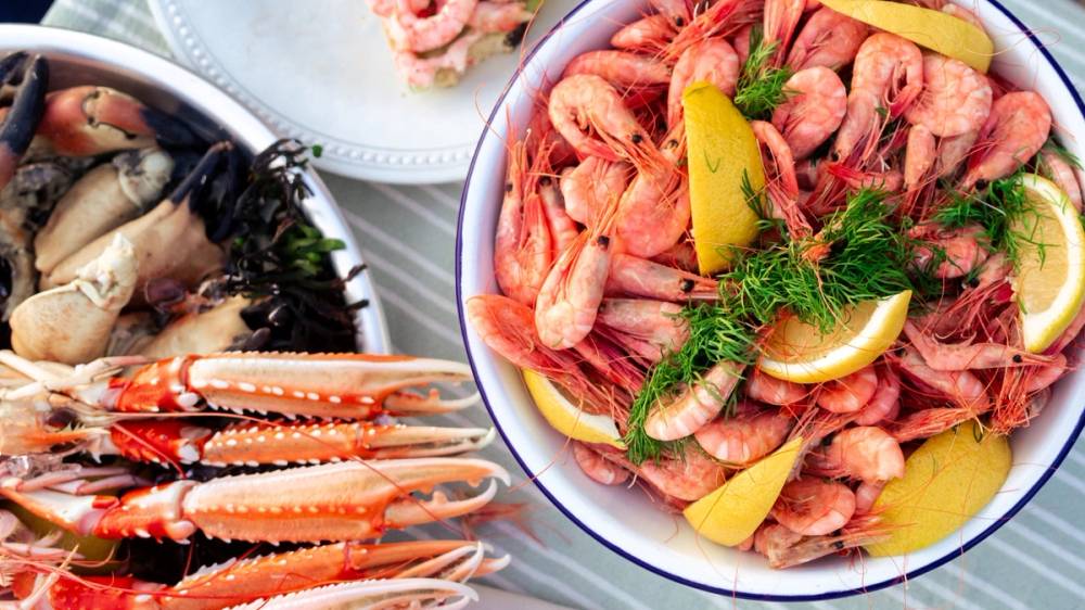 En bolle med reker, sitronbåter og dill, omgitt av sjømat og tilbehør på et dekket bord.