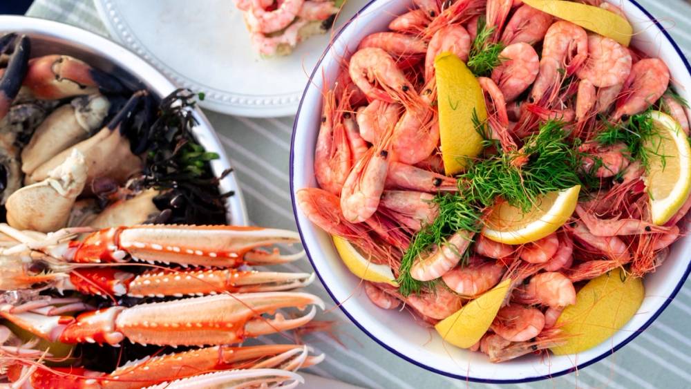 En bolle med reker, sitronbåter og dill, omgitt av sjømat og tilbehør på et dekket bord.