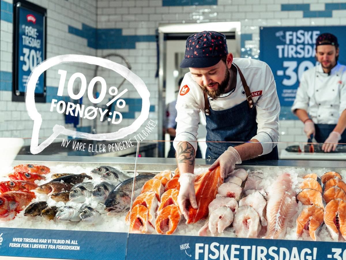 En person i kjøkkenuniform som skjærer opp fisk i en fiskebutikk, med andre ansatte i bakgrunnen.