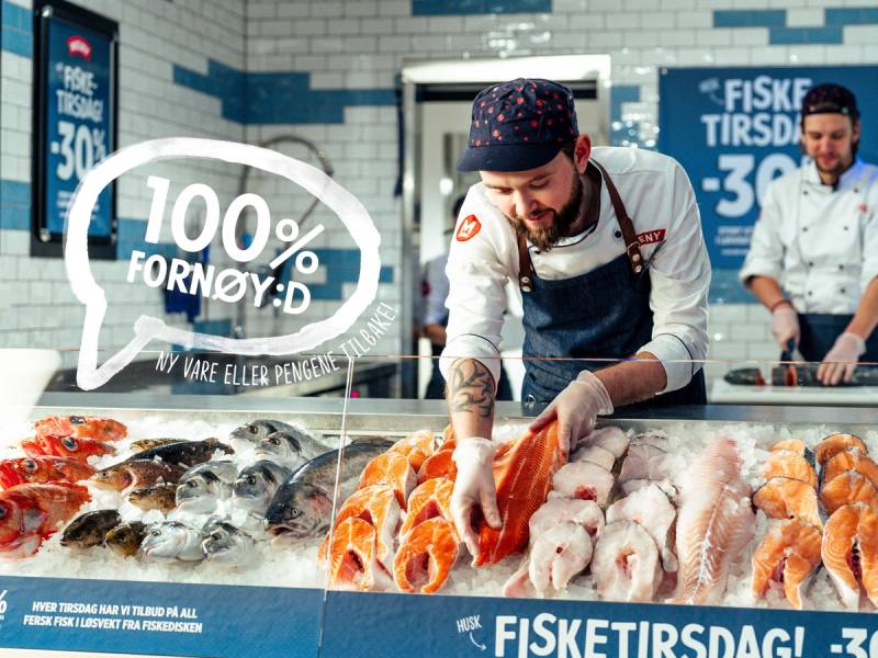 En person i kjøkkenuniform som skjærer opp fisk i en fiskebutikk, med andre ansatte i bakgrunnen.