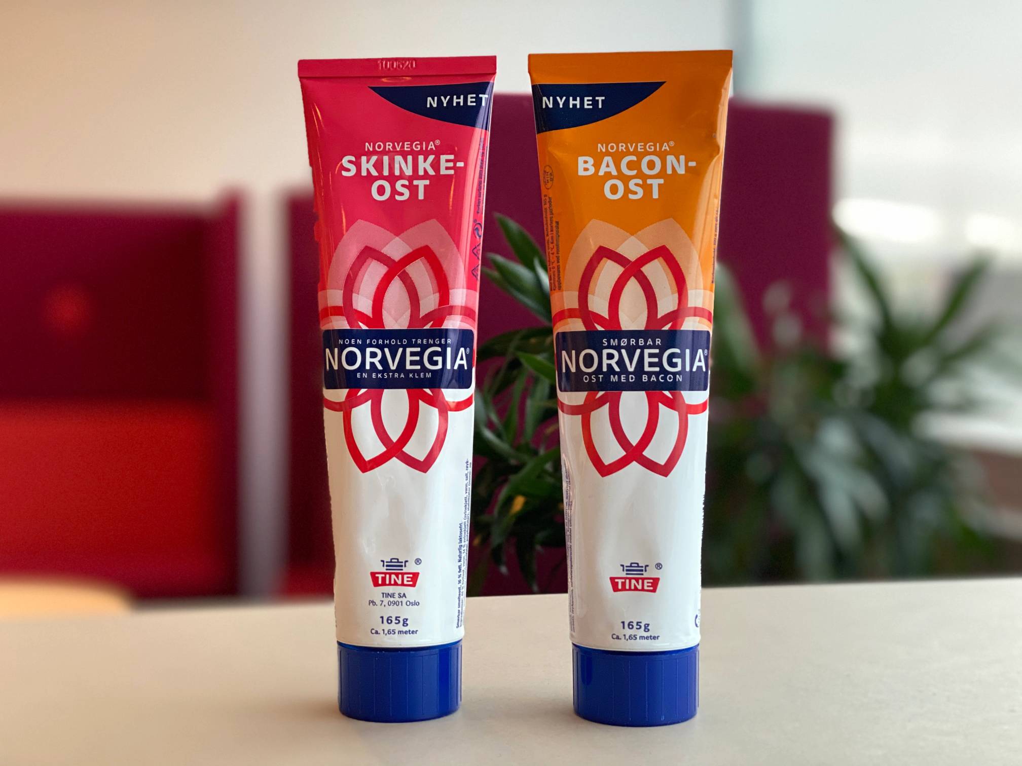 To tuber med Norvegia skinkeost og baconost på et bord med en bakgrunn av grønne planter og røde stoler.