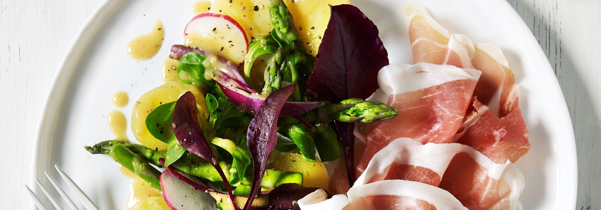 Tallerken med skiver av spekeskinke og en frisk salat med grønnsaker og urter.