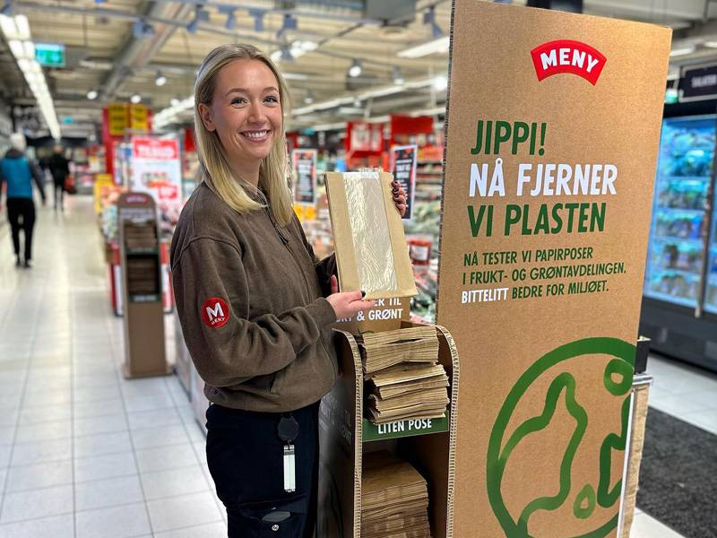 En person i butikk som kaster plastemballasje i en resirkuleringsstasjon for plast, med en stor informasjonsplakat om plastgjenvinning ved siden av.