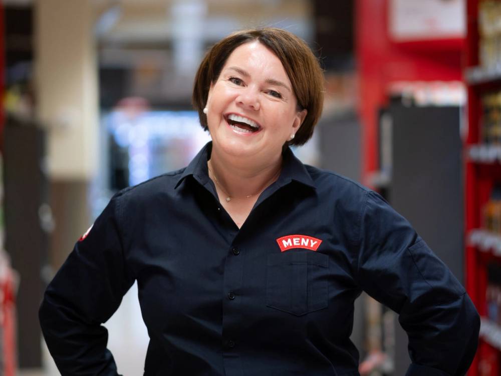 En person med kort hår iført en mørk uniform med en Meny-logo på brystet, står i en butikk eller lagerhall.