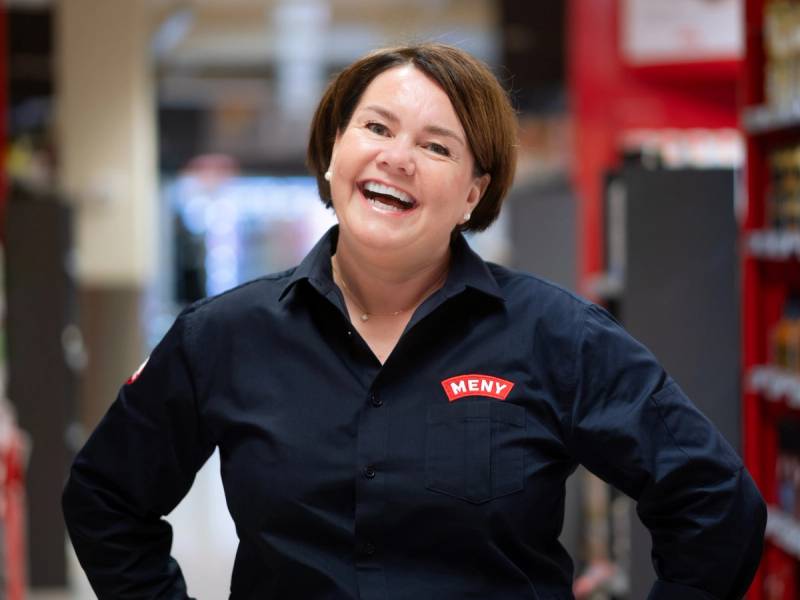 En person med kort hår iført en mørk uniform med en Meny-logo på brystet, står i en butikk eller lagerhall.