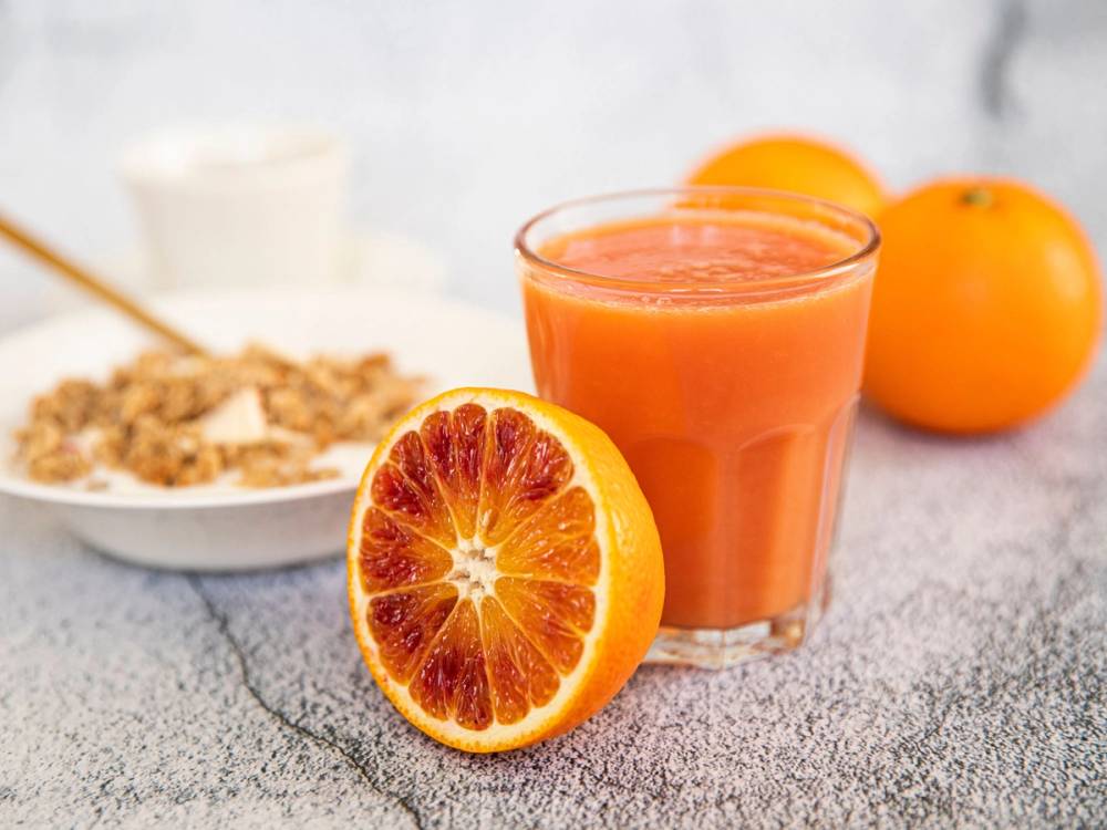 Et glass med blodappelsinjuice står foran en halvert blodappelsin og to hele appelsiner på et kjøkkenbord.