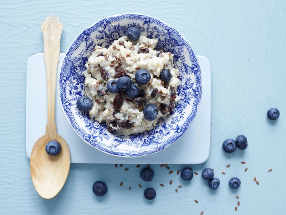 En skål med enkel havregrøt toppet med blåbær og rosiner, plassert på en hvit skjærefjøl med en tresleiv ved siden av.