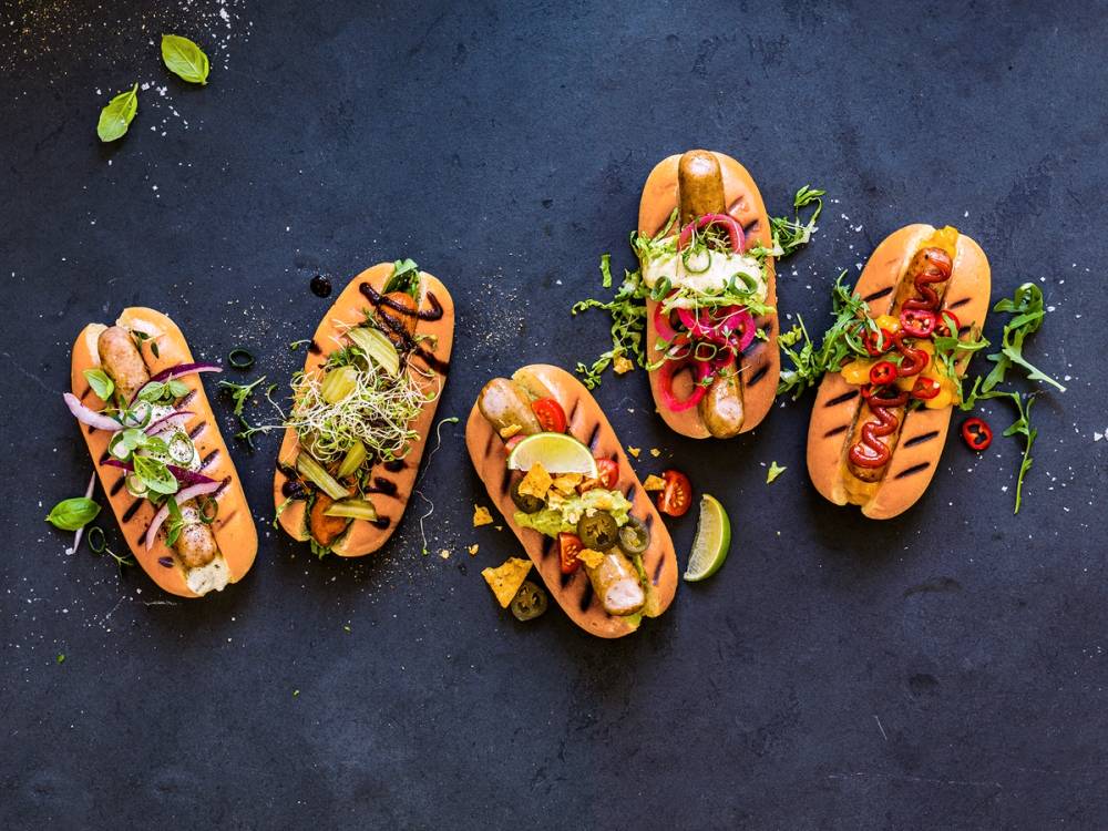 Fem pølser med ulike pålegg og garnityr på en mørk bakgrunn, pyntet med urter, grønnsaker og saus.