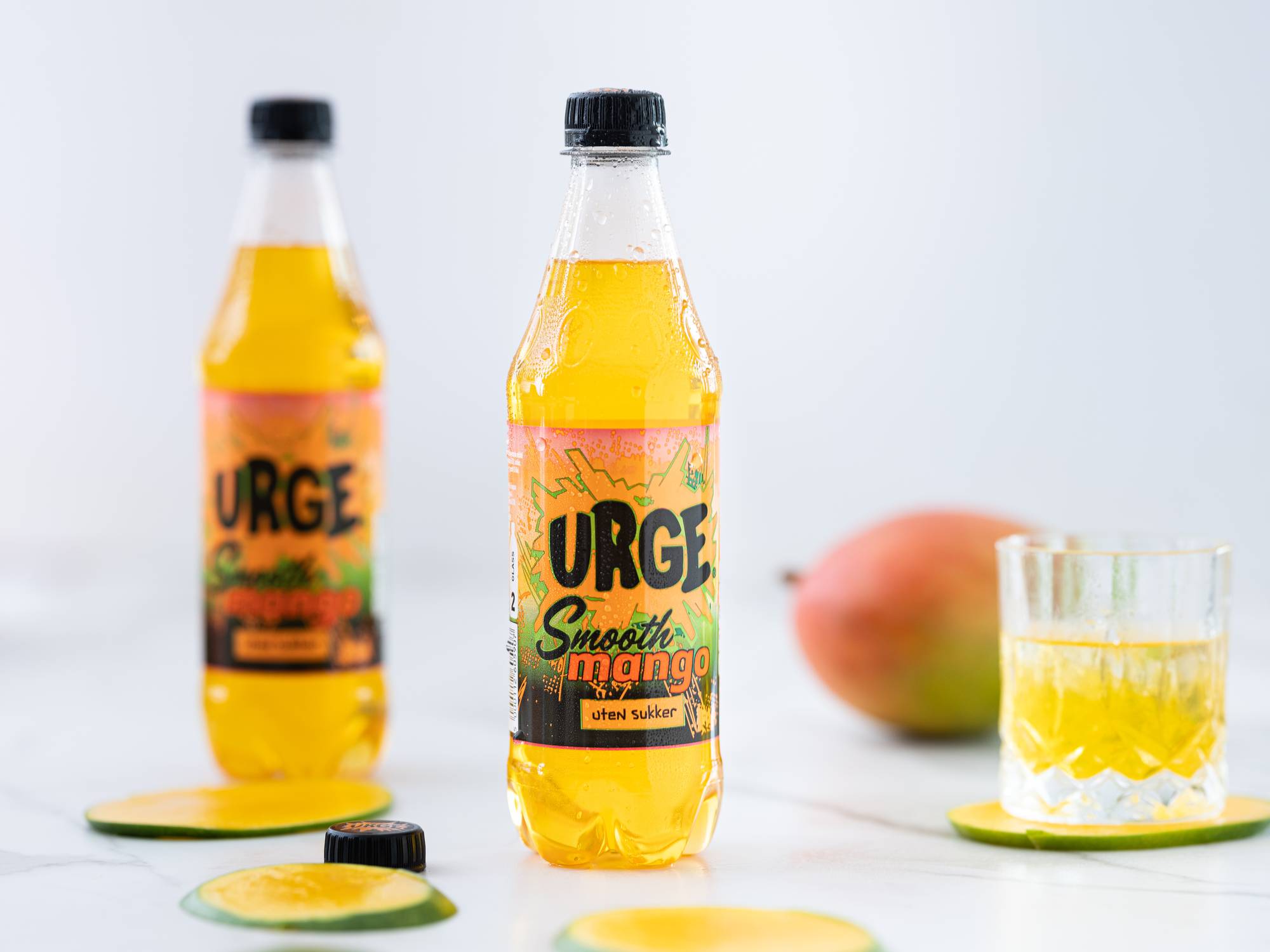 To flasker med URGE Smooth mango uten sukker og et glass med mango-smoothie på et bord med mango og sitron i bakgrunnen.