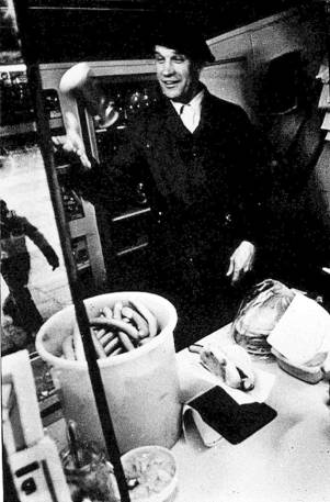 En person i mørk dress står bak en butikkdisk med ulike varer, inkludert en stor beholder med pølser, i en butikk eller kafé.