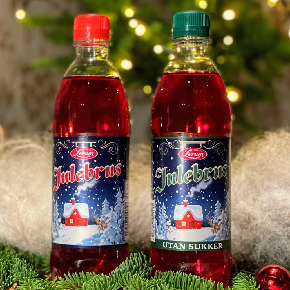 To flasker med Lerum julebrus, en med rød hette og en med grønn hette, står på et bord med juledekorasjoner foran et juletrær med lys i bakgrunnen.