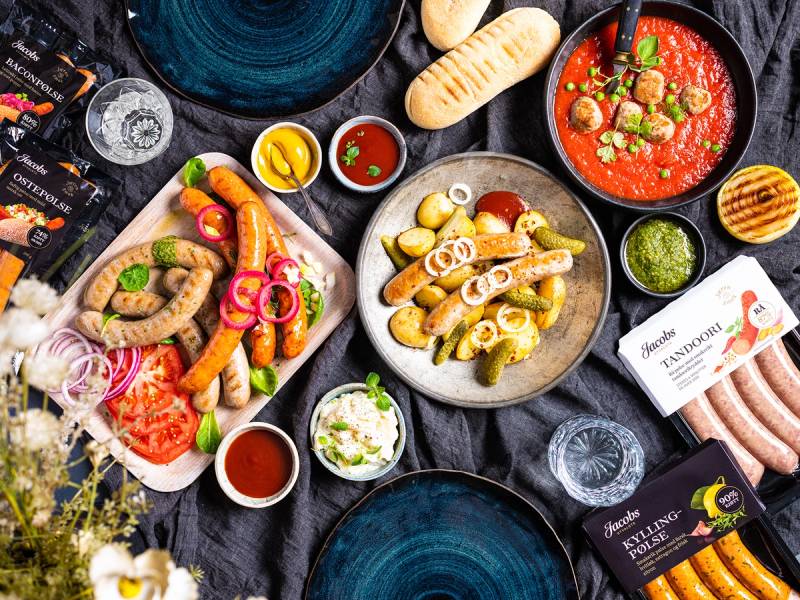 Et utvalg av ulike matretter og tilbehør på et bord, inkludert pølser, brød, sauser og grønnsaker.