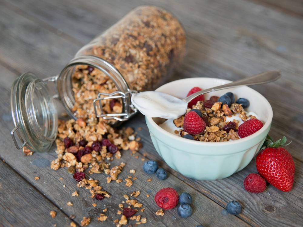 En bol med granola, friske bær og yoghurt, med en glasskrukke med granola ved siden av på et trebord.