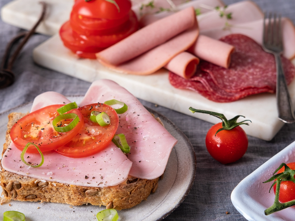 En skive med pålegg med skinke, toppet med tomatskiver og grønn løk, med flere typer pålegg og tomater i bakgrunnen.