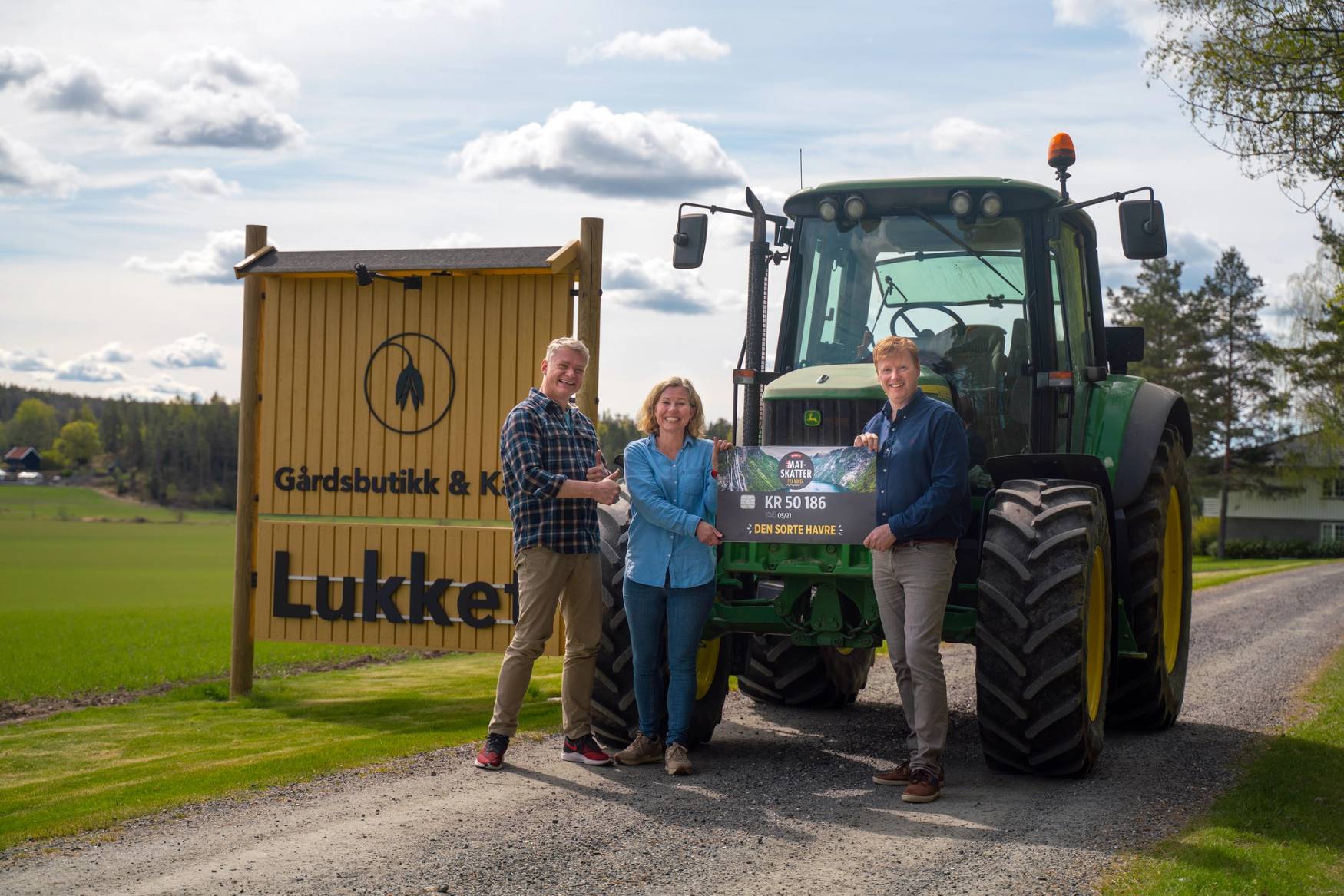 Tre personer står foran en stor grønn traktor og et skilt som sier 'Lukket', på en grønn gresslette med trær og en blå himmel i bakgrunnen.