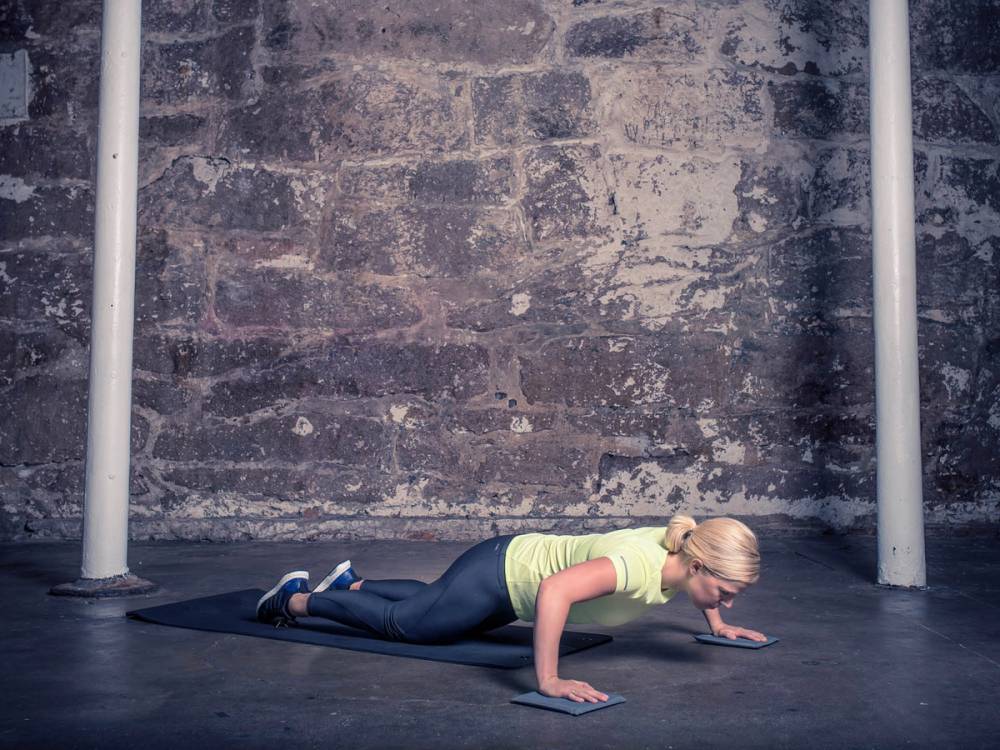 En person gjør en push-up-øvelse på gulvet foran en murvegg, med to hvite stenger på hver side.