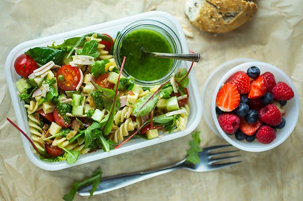 En matpakke med grønn salat, en liten bolle med bær, og en glass med grønn dressing, på et lyst bord.