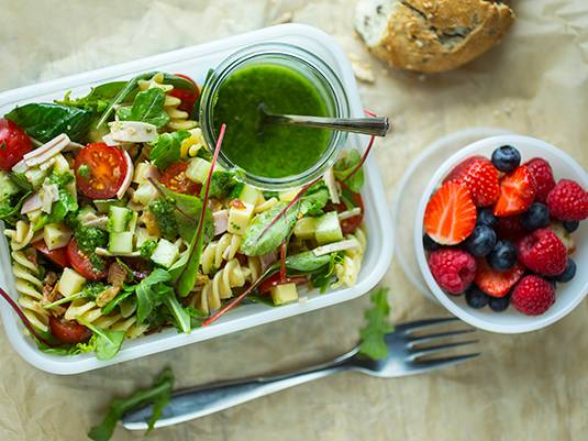 En matpakke med grønn salat, en liten bolle med bær, og en glass med grønn dressing, på et lyst bord.