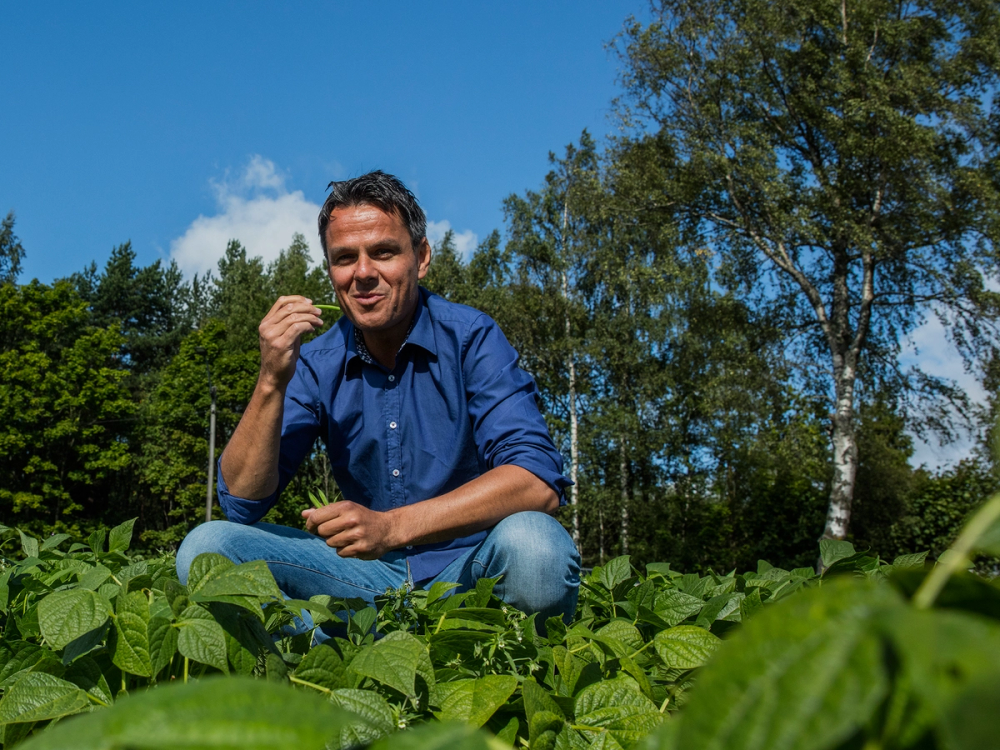 En person i blå skjorte som sitter blant grønne planter i en hage eller frukthage med trær i bakgrunnen på en solrik dag.
