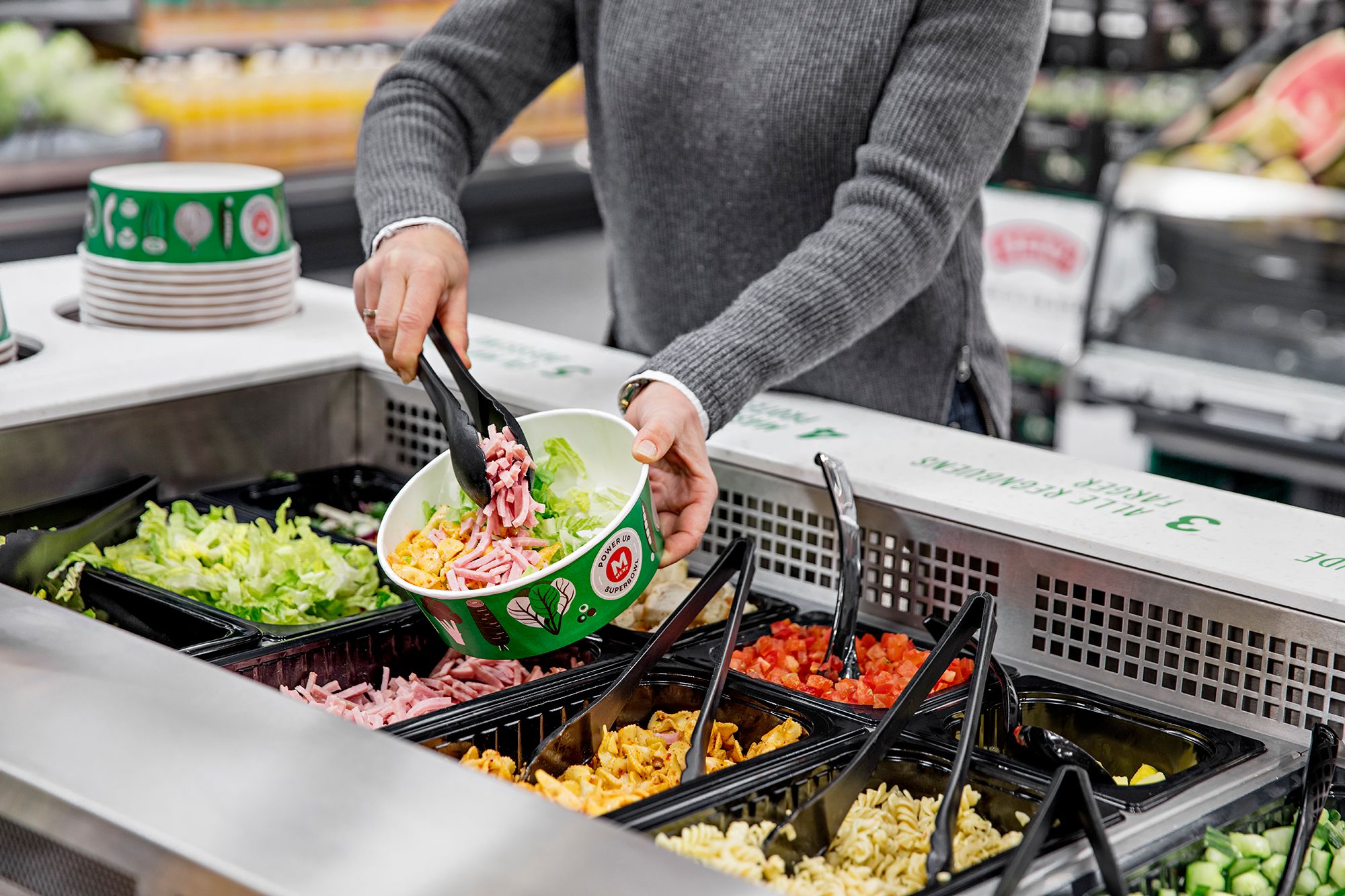 En person plukker salat fra en salatbar med ulike grønnsaker i beholdere.