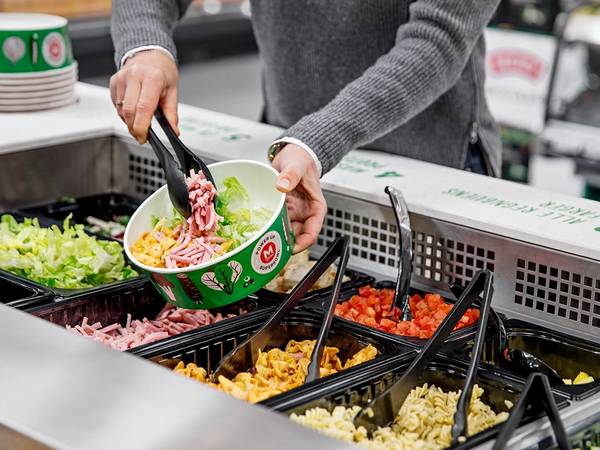 En person plukker salat fra en salatbar med ulike grønnsaker i beholdere.